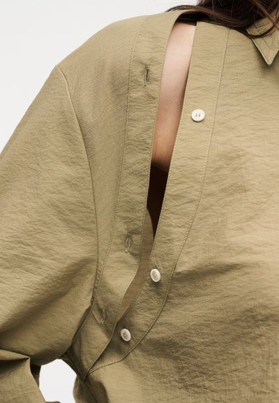 Κοντινό πλάνο μπεζ πουκάμισου με κουμπιά και ανοιχτή πλευρική ραφή που αποκαλύπτει το δέρμα από κάτω σε ένα άτομο.