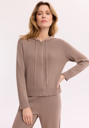 Kobieta z jasnobrązowymi włosami, ubrana w taupe'ową bluzę rozpinaną na suwak i pasujące spodnie, stojąca z jedną ręką na biodrze na jednolitym tle.