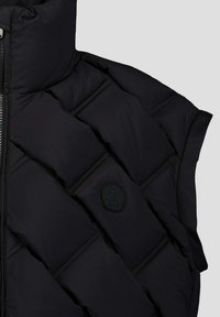 Zwarte gewatteerde jas mouw met horizontale stiksels en een zwart rond JOTT-logo patch, weergegeven tegen een lichtgrijze achtergrond.