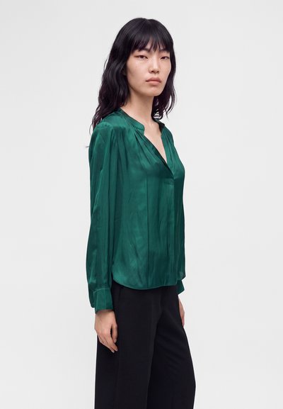 Femme aux longs cheveux noirs portant un chemisier vert brillant à manches longues et un pantalon noir, debout devant un fond blanc uni.