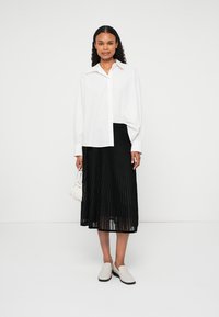 Cămașă albă cu nasturi, fustă midi plisată neagră, pantofi deschiși, ținând o geantă mică albă. Design simplu cu țesătură fluidă și linii clare.