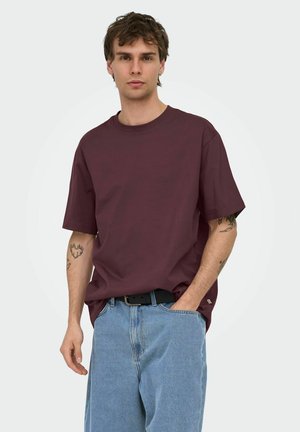 Jeune homme portant un t-shirt uni bordeaux foncé avec un jean bleu clair, une main dans la poche, tatouages visibles sur les deux avant-bras, fond neutre.