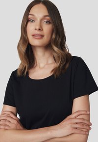 Czarna bluzka z krótkim rękawem i okrągłym dekoltem, wykonana z miękkiego materiału. Modelka ma falowane włosy do ramion, ręce skrzyżowane na piersiach.