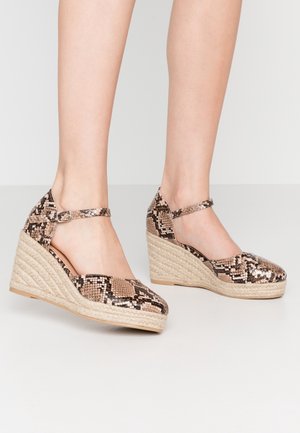 Nogi w beżowo-brązowych koturnach ze wzorem skóry węża, z paskami wokół kostek, na białej podłodze na tle jasnego, jednolitego tła.