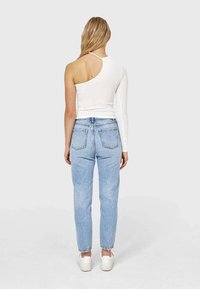Mujer con cabello largo y rubio que lleva una blusa blanca asimétrica con una manga larga, jeans azul claros y zapatillas blancas, de pie de espaldas.