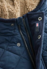 Navy quilted jacket with shearling lining, zipper, snap buttons, ribbed collar, and a label showing a UK flag and brand details.