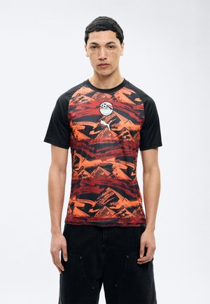 Νεαρός άνδρας με κοντά σγουρά μαλλιά, ντυμένος με αθλητική φανέλα κόκκινου και μαύρου μοτίβου με μαύρα μανίκια και μαύρο παντελόνι, στέκεται μπροστά σε λευκό φόντο.