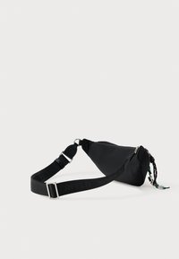 Černá nylonová ramenní taška s trojúhelníkovým tvarem, nastavitelný popruh s vyraženým logem a zipové uzavírání se dekoračními kovovými prvky.