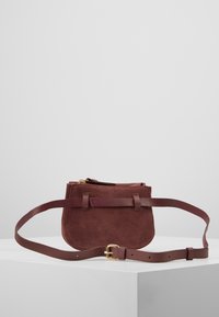 Skarpetek burgundowy z zamszu, z zamkiem błyskawicznym, o płaskim kształcie, z regulowanym skórzanym paskiem i dwoma skórzanymi akcentami z przodu.