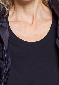 Svart ärmlös topp med rund halsringning, bärs under en mörk puffjacka. Tyget verkar slätt och åtsittande.