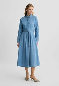 Helles, blaues Denim-Kleid mit durchgehender Knopfleiste, langen Ärmeln und einem Gürtel in der Taille. Ausgestellter, knielanger Rock und ein Kragen-Detail.