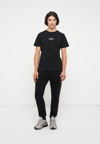 Camiseta negra de algodón con un pequeño logotipo gráfico en el pecho, combinada con pantalones de chándal negros que tienen una raya lateral roja. Llevada con zapatillas grises.
