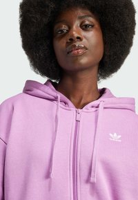 Levandulová mikina na zip z měkkého materiálu s přední kapsou, šňůrkami na kapuci a malým bílým logem Adidas na levém prsu.
