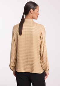 Beżowa bluzka w pionowe paski, wykonana z lekkiego materiału, z długimi rękawami, luźny fason i wysoki kołnierz; gładka faktura z delikatnym połyskiem.