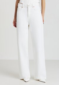 Persona che indossa jeans bianchi a vita alta e a gamba larga con cinque tasche e scarpe bianche con punta a punta, in piedi davanti a uno sfondo neutro.