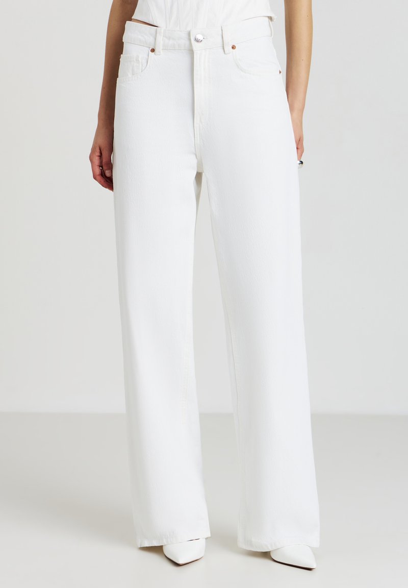 Persona che indossa jeans bianchi a vita alta e a gamba larga con cinque tasche e scarpe bianche con punta a punta, in piedi davanti a uno sfondo neutro.