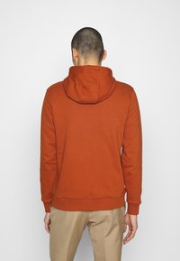 Felpa arancione in cotone con tasca posteriore e cappuccio regolabile. Tessuto liscio, polsini e vita a coste. Indossata con pantaloni beige.