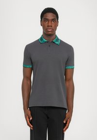 Mørk grå polo skjorte med korte ermer, med en krage og ermekanter i turkise mønstre. Glatt tekstur og klassisk passform.