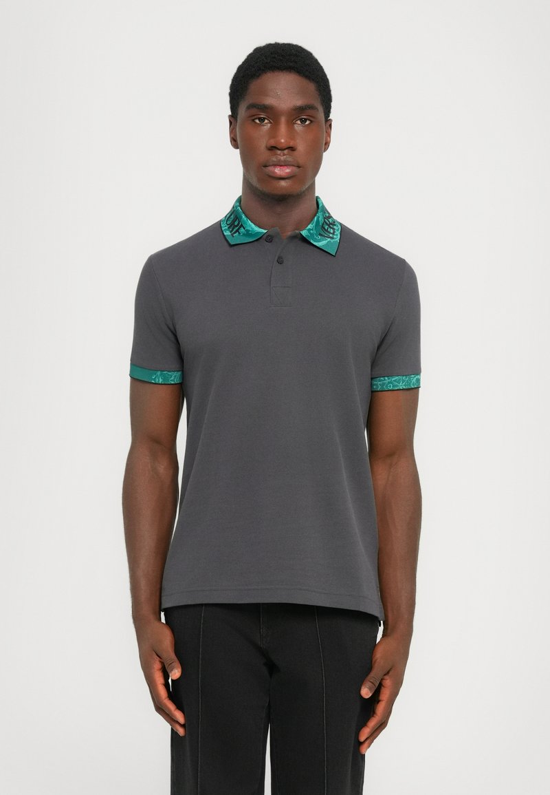 Mørk grå polo skjorte med korte ermer, med en krage og ermekanter i turkise mønstre. Glatt tekstur og klassisk passform.