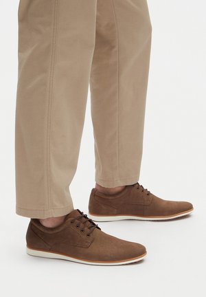 Brown canvas shoes with brown leather accents, featuring a round toe, lace-up design, and a white rubber sole, paired with beige trousers.