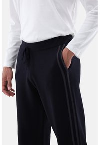 Person in schwarzen Jogginghosen mit Kordelzug und Seitennstreifen, mit einer Hand in der Tasche, kombiniert mit einem weißen Langarmshirt.