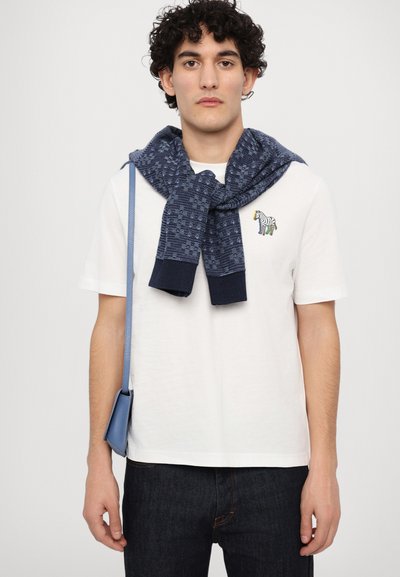 Balta kokvilnas T-krekls ar krāsainu zebras izšuvumu, papildināts ar tumši zilu teksturētu adījuma džemperi, kas uzlikts uz pleciem, un zilu plecu somu.