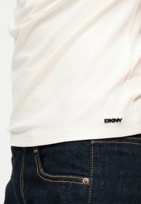 Κοντινή λήψη λευκής ραφής πουκάμισου DKNY πάνω από σκούρα μπλε τζιν με ορατό πορτοκαλί ράψιμο και μεταλλικές λεπτομέρειες με λάμες.
