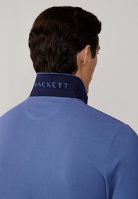 Cuello azul marino con "HACKETT" en texto azul claro. La camisa es de un azul claro sólido, hecha de una tela suave y con un corte relajado.