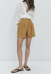 Leinenshorts in einem senfgelben Farbton mit vorderen Falten und Knopfverschluss, kombiniert mit einem lockeren weißen Hemd mit Knöpfen und schwarzen Flip-Flops.