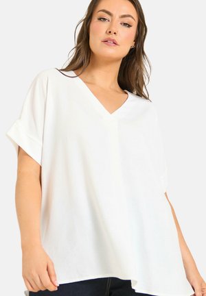 Kvinde med langt brunt hår iført en løs hvid V-hals kortærmet top og mørke bukser, poserer mod en ensartet lys baggrund.