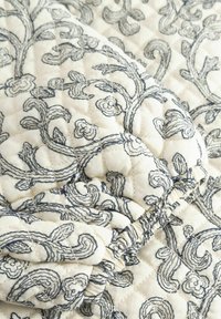 Tkanina pikowana z misterną niebieską haftowaną kwiatową dekoracją na kremowym tle, o miękkiej i strukturalnej powierzchni.