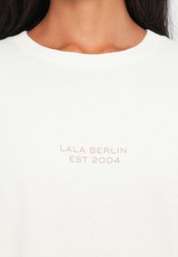 Biała bluza z okrągłym dekoltem; nadrukowany tekst „LALA BERLIN EST 2004” w miękkim beżowym kolorze, umieszczony na środku klatki piersiowej.