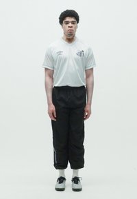 Balts īsām piedurknēm jersejs ar melnām svītrām un logo, apvienots ar melnām sporta biksēm un baltām zeķēm. Modelis stāv uz vienkārša fona.