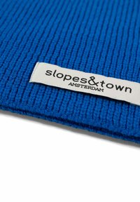 Un textile bleu à côtes avec une étiquette cousue portant l'inscription "slopes&town AMSTERDAM" en texte noir. Le matériau semble tricoté et texturé.