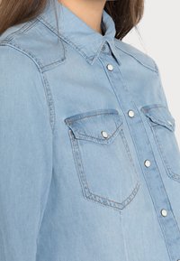 Ljust blå denimskjorta med strukturerad krage, tryckknappar och två bröstfickor. Har kontrasterande sömmar för extra detalj.