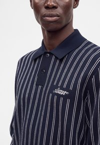 Homme portant un polo bleu marine avec des fines rayures verticales blanches et un logo brodé sur la poitrine gauche.