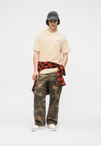 Νεαρός που φοράει μπεζ μπλουζάκι McLaren, παντελόνι καμουφλάζ, λευκά αθλητικά παπούτσια, μαύρο καπέλο και κόκκινο-μαύρο καρό πουκάμισο δεμένο γύρω από τη μέση.