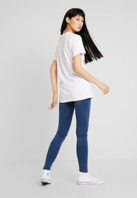 Weißes Baumwoll-T-Shirt mit kurzen Ärmeln, kombiniert mit blauen Skinny-Jeans und weißen High-Top-Sneakern. Das Model posiert seitlich.