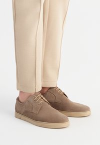 Hellbraune Wildlederschuhe mit runder Zehenform, Schnürdesign und Gummisohle. Kombiniert mit hellbeigen, leicht taillierten Hosen.