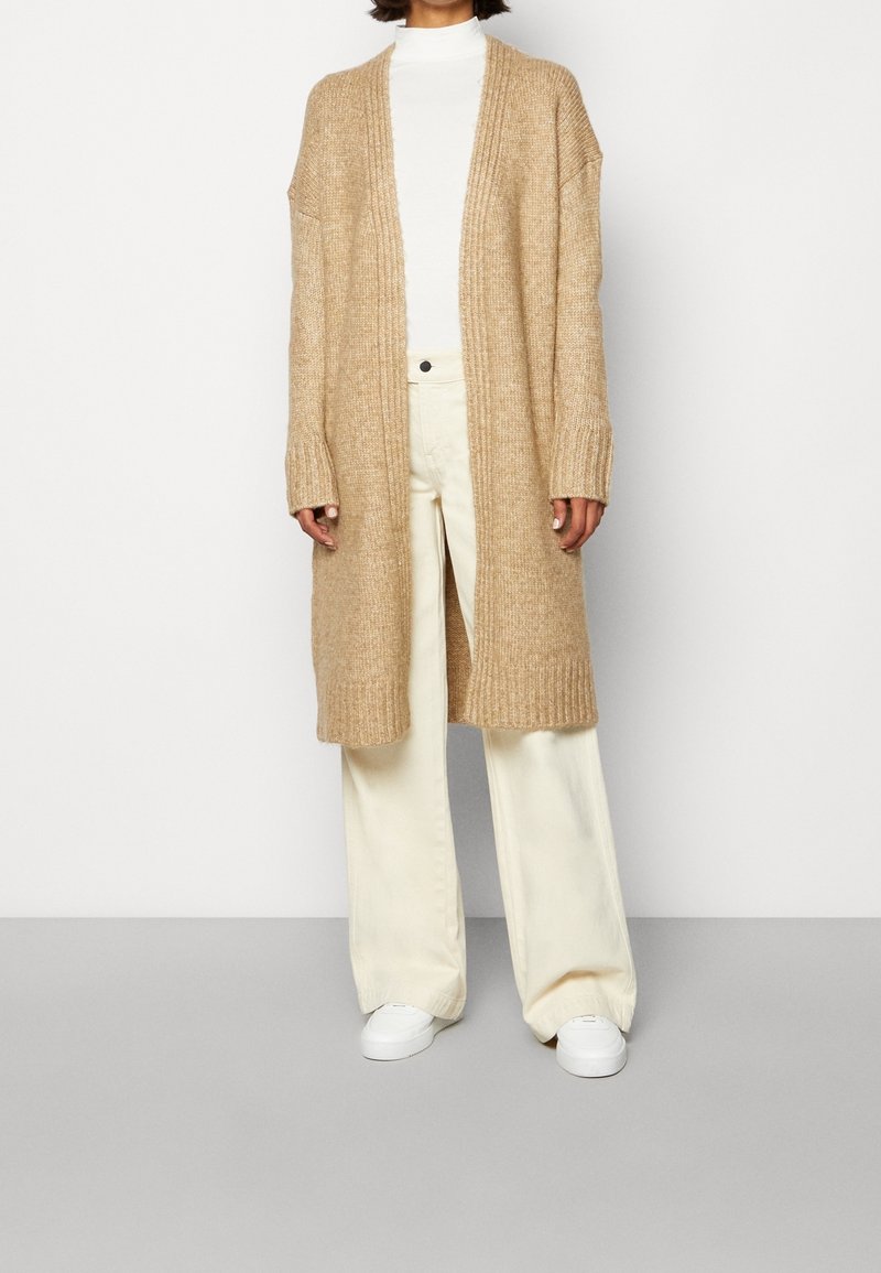 Personne portant un long cardigan beige en tricot par-dessus un col roulé blanc, un pantalon large crème et des baskets blanches, debout devant un fond uni.