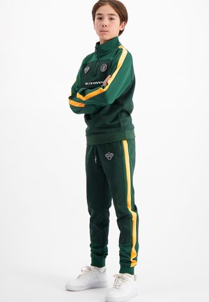 Groen sporttrainingspak met gele zijstrepen, een jasje met rits en bijpassende taps toelopende broek. Witte sneakers maken de outfit compleet.