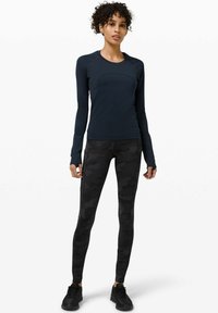 Langärmliges navyblaues Sportoberteil, kombiniert mit schwarzen Camouflage-Leggings aus dehnbarem Material und Daumenlöchern an den Ärmeln. Schwarze Sneaker.