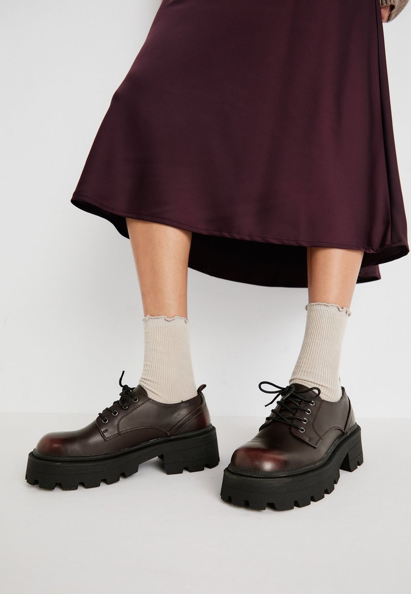 Scarpe platform in pelle marrone con suole nere spesse, abbinate a calzini beige a coste con bordi a scallop, su uno sfondo tenue.