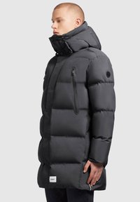 Mustan puffjakiga, millel on kõrge krae, lukuga ja nööbiga sulgemine, tekstureeritud paneelid ja logo silt alläärs. Külgmine tõmblukuga tasku on nähtav.