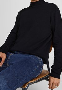 Pull noir en maille avec un col montant, des poignets côtelés et une texture douce, assorti à un jean en denim bleu, assis sur une chaise en bois.