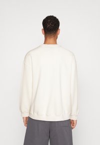 Uomo dai corti capelli ricci, che indossa una felpa bianco sporco e pantaloni grigi, in piedi di spalle su uno sfondo leggero e uniforme.
