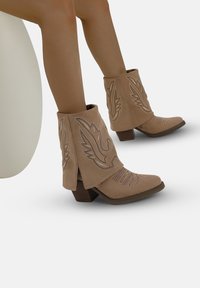 Botas de cowboy beige con un acabado de ante texturizado, acentos de costura decorativa y tacones en bloque angulados. Las botas presentan un diseño holgado.