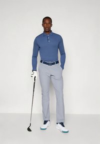 Roupa de golfe composta por uma polo de manga comprida azul-marinho, calças cinza claro e sapatos brancos com detalhes azuis. Segurando um taco de golfe com uma luva branca.