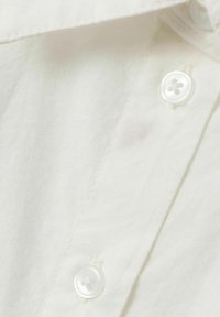 Camicia bianca in cotone leggero con una superficie liscia, dotata di cinque bottoni bianchi e sottili macchie rosse vicino al collo.