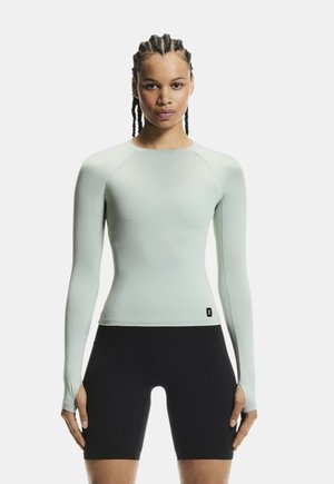 Femme aux cheveux nattés portant un haut ajusté à manches longues gris clair avec des trous pour les pouces et un short de cyclisme noir, debout devant un fond blanc.